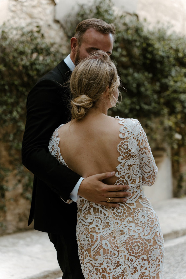 Braut in Boho Brautkleid mit Spizte und lockerer niedrig gesteckter Frisur steht neben Bräutigam mit dem Rücken zur Kamera