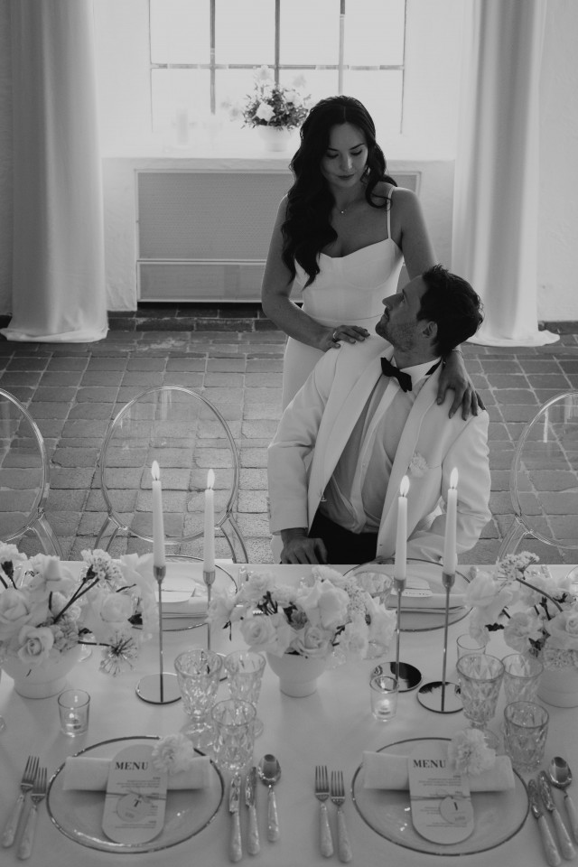 Braut steht hinter dem Bräutigam welcher auf einem Stuhl am Hochzeitstisch sitzt