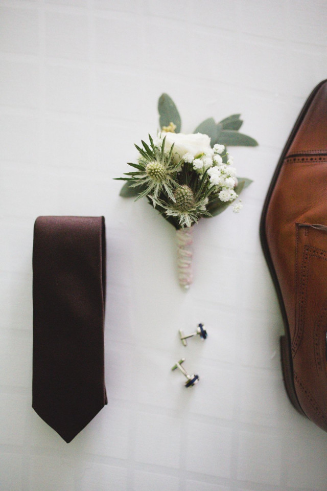 Flat lay met een donkere stropdas, een witte boutonnière, manchetknopen en een lederen schoen.