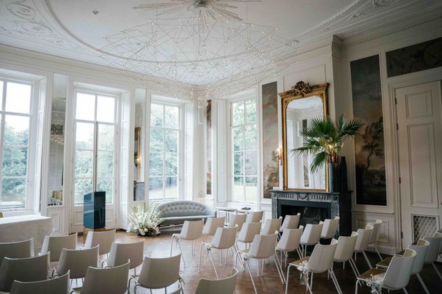 Lege ceremoniezaal met rijen witte stoelen, hoge ramen, een grijze bank en een grote vergulde spiegel boven de schouw.