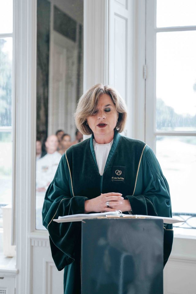 An officiant in a green robe speaks behind a lectern by tall windows during the ceremony.