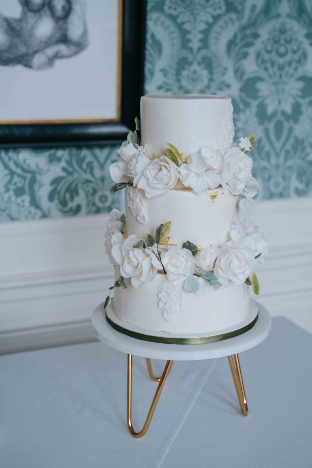A three-tier white wedding cake decorated with white roses on a gold stand.