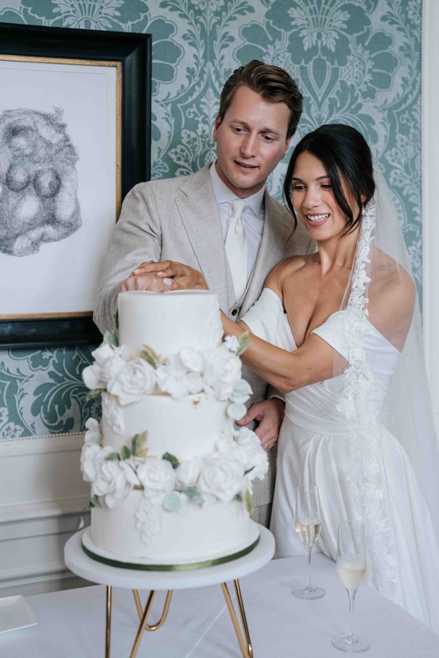 A couple cut a white wedding cake decorated with roses together beside a framed photo.