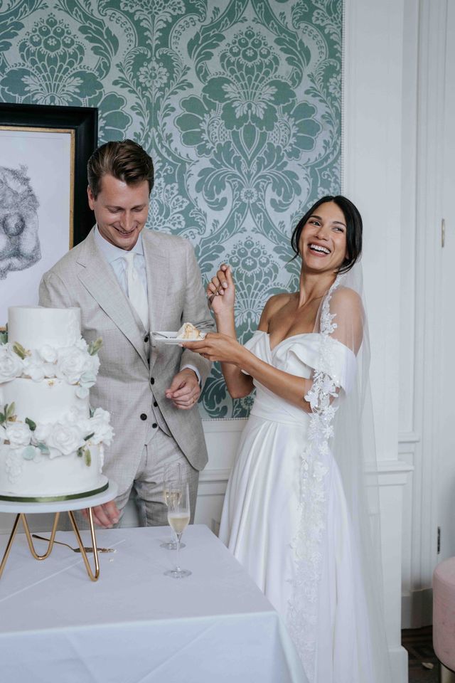 A smiling couple stand beside their white wedding cake on a gold stand at the reception.