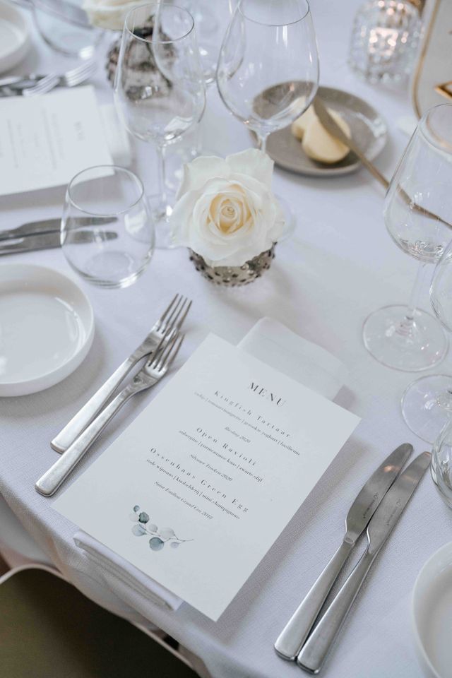 A place setting with a printed menu card, a single white rose, and polished silver cutlery.