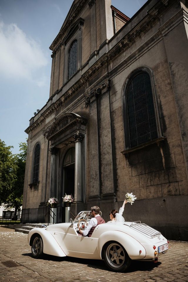 A bride and groom ride in a white vintage convertible past a classic building facade.