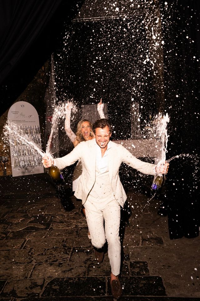 A couple in light suits run laughing through sparklers and champagne at their wedding.