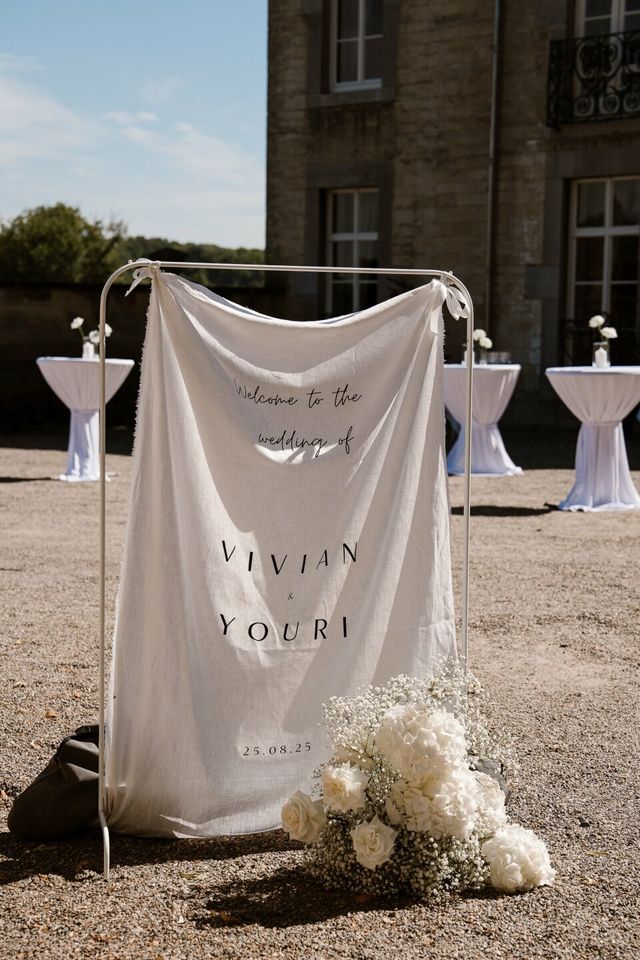 A linen welcome banner reads 'Welcome to the wedding' beside a white floral arrangement.