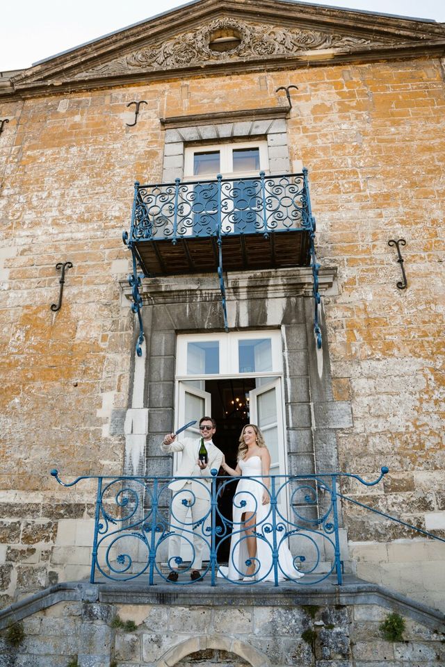 A couple stand with a champagne bottle on a blue wrought-iron balcony in the morning light.
