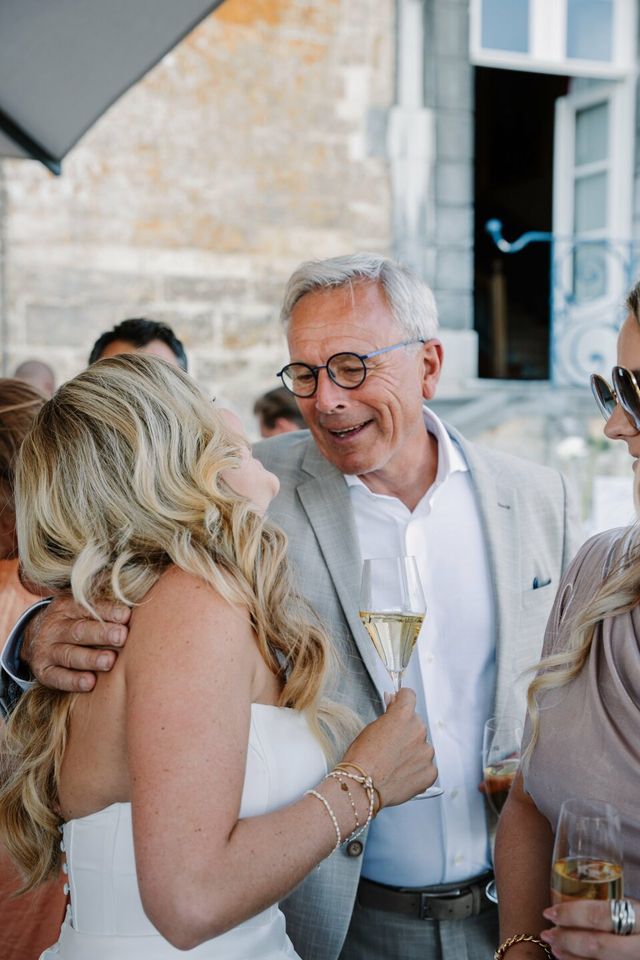 The bride embraces a smiling older guest, champagne in hand, at the reception.
