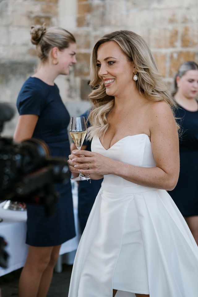 The bride in a short white dress laughs while holding a glass of champagne.