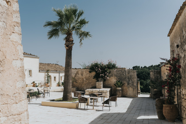 A sunlit whitewashed masseria courtyard with a palm tree, stone walls, and cafe tables.