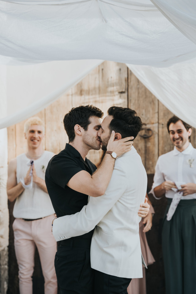Two grooms share a kiss under a soft white drapery at the ceremony.