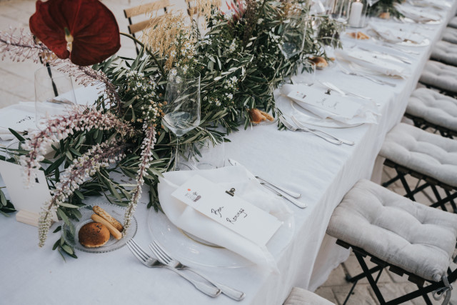 Detail of a long set table with a white runner, green branches, and red anthurium.