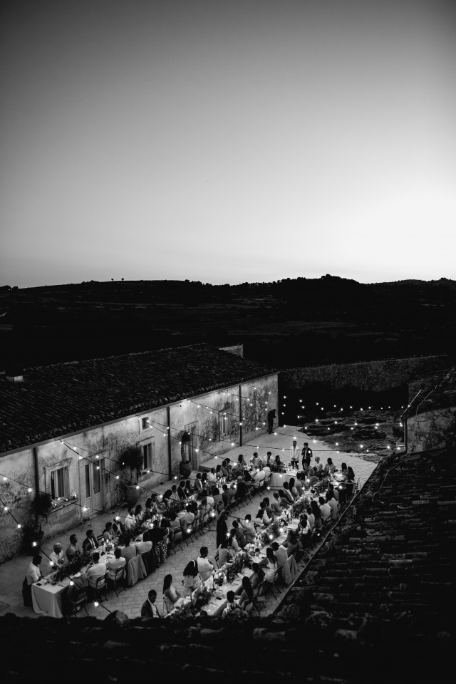 A black and white overview of an evening dinner on a lit courtyard at a wedding venue.
