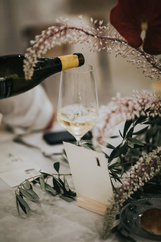 Detailfoto van witte wijn die in een glas wordt geschonken naast roze bloementakken en een naamkaartje op tafel.