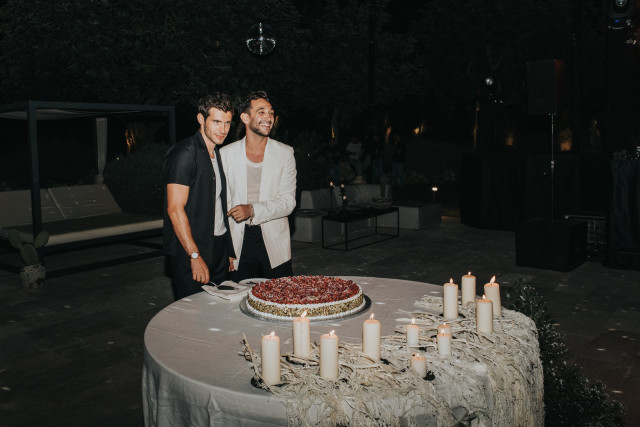 Two guests stand by a round cake on an outdoor table at the celebration.