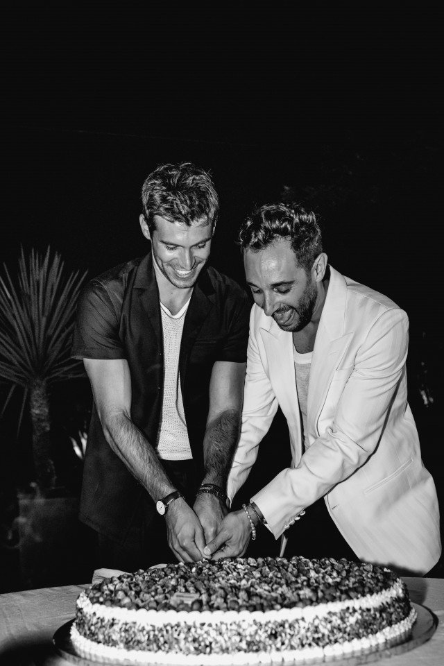A black and white photo of two smiling grooms cutting their wedding cake together.