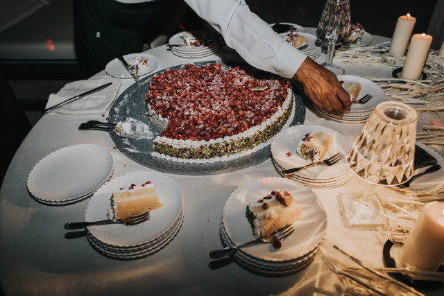 A hand slices a large round cake topped with red fruit at the reception.