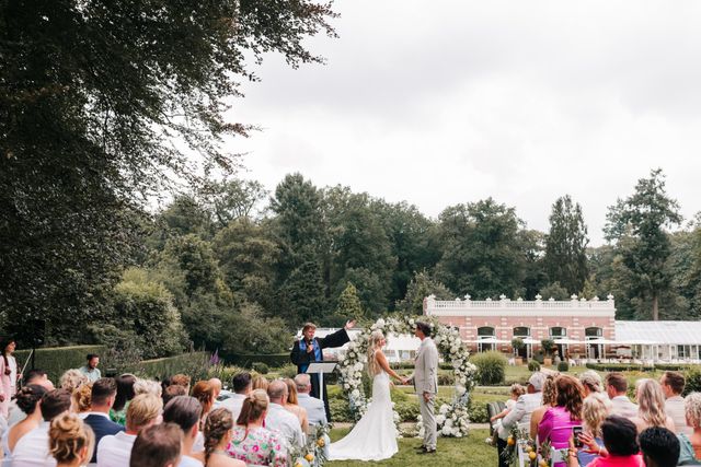 Overzichtsopname van een buitentrouwerij in een tuin met zittende gasten en het paar dat de geloften uitspreekt bij een bloemenboog.