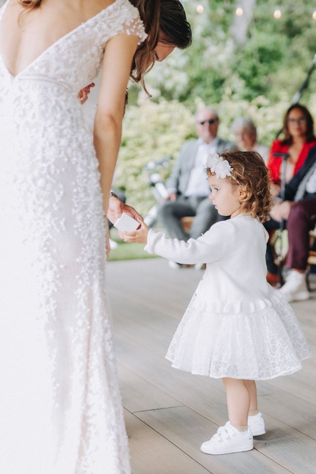 An adorable flower girl in a white dress and flower headband holds hands with the bride during the ceremony.