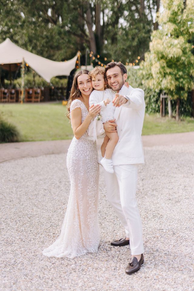 The happy couple poses outdoors with their young child in arms, all three smiling, the wedding tent behind them.