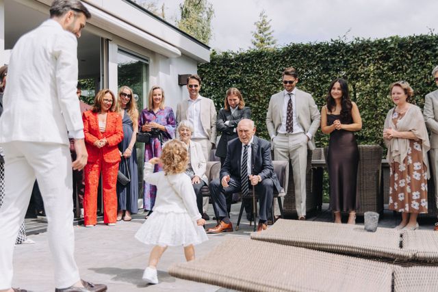 A young girl in a white dress runs playfully toward the groom outdoors, delighting nearby guests.