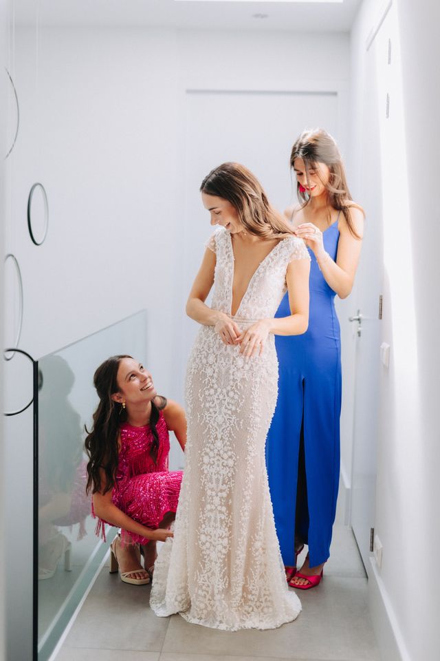 A bride smiles as two bridesmaids help her into her lace wedding gown.