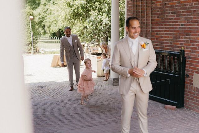A flower girl in a pink dress runs playfully between two groomsmen in beige suits outside the wedding venue.