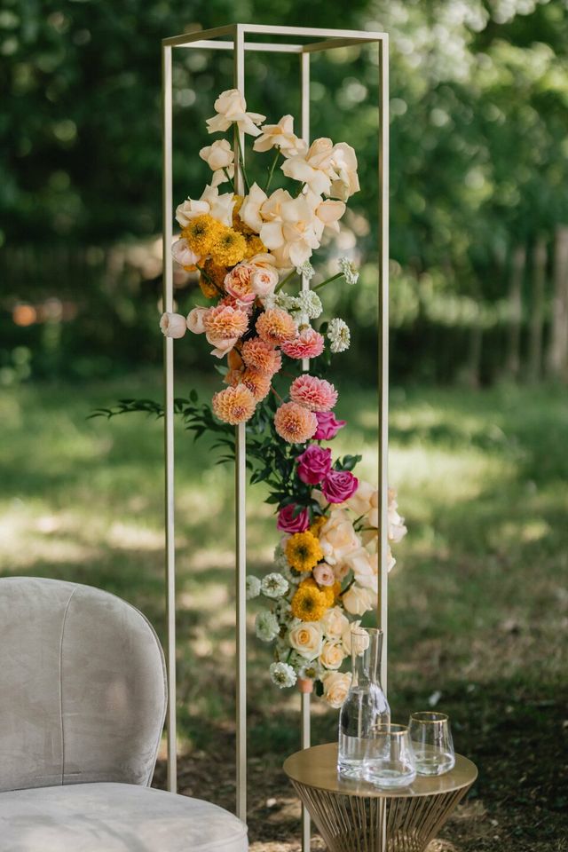 A tall gold frame floral installation bursting with pink, yellow, peach, and white blooms in a garden.