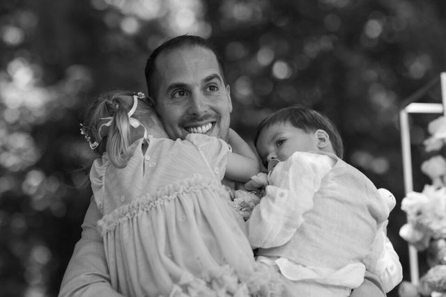 Black and white photo of the groom holding two young children, all three smiling, at the outdoor ceremony.