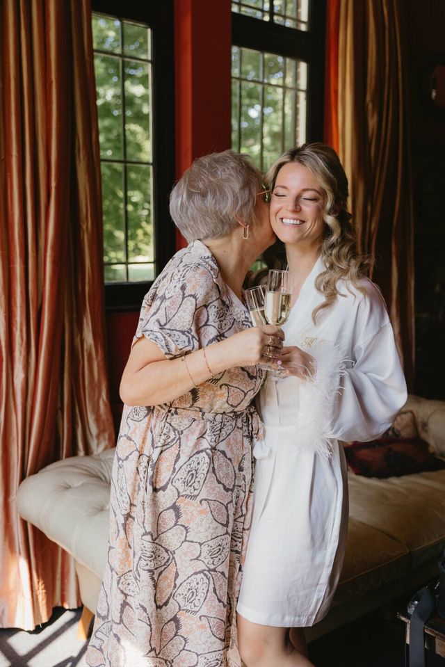 A bride in a white robe and her mother share a joyful champagne toast during morning wedding preparations.