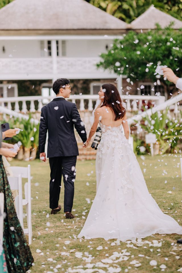 A couple walk away hand in hand as white flower petals shower down around them, a tropical estate behind.