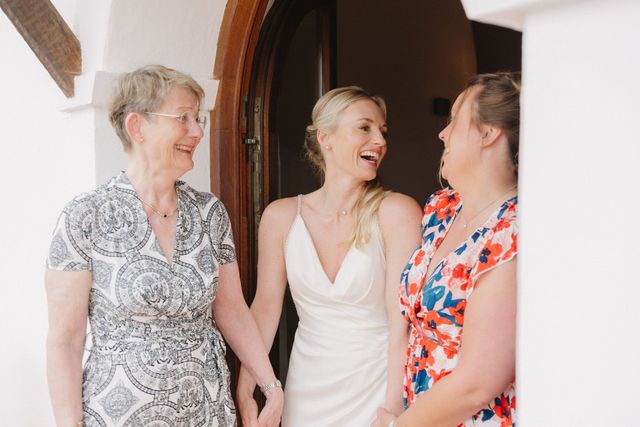 A bride laughs warmly with her mother and a close family member in a sunny doorway before the ceremony.