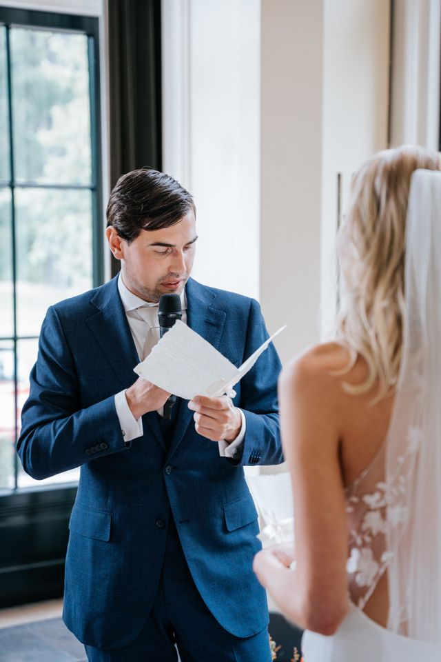 A groom in a dark blue suit reads his vows carefully from a paper during the ceremony.