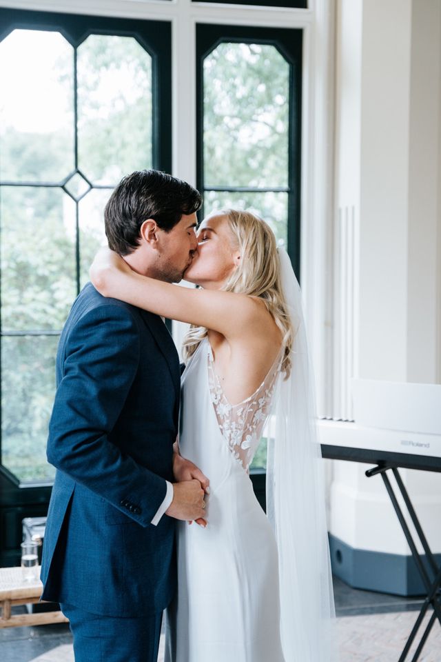 The couple kisses while standing in front of tall windows at the venue.