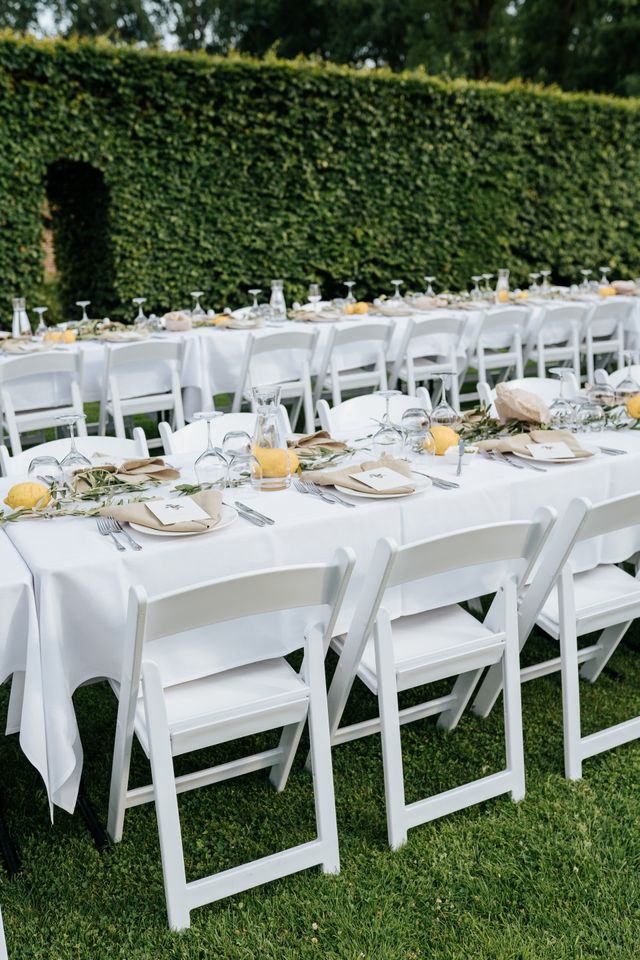 Long white-set party tables with white folding chairs arranged on the grass.