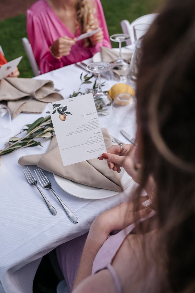 A guest reads a printed menu card with a citrus illustration above a white set table.