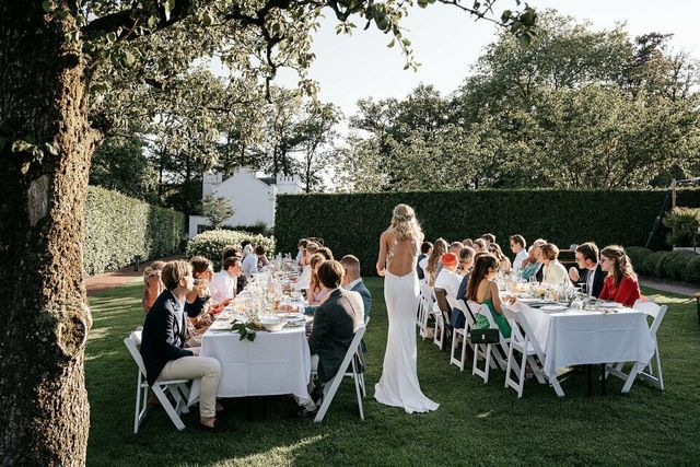 Bruid loopt naar lange gedekte dinertafels vol gasten in een zonnige tuin tijdens het bruiloftsdiner.