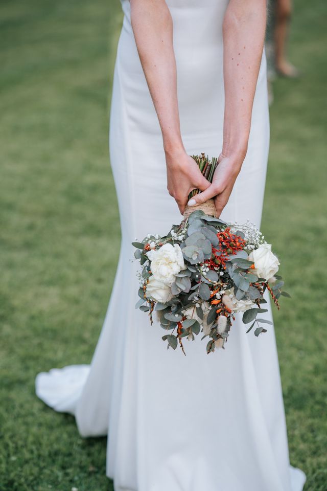 A bride holds a bouquet of eucalyptus and white roses against her dress.