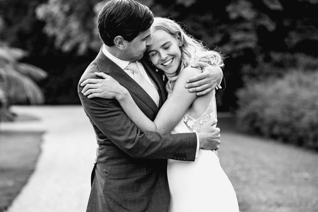 A couple embraces on a garden path, the bride smiling with her eyes closed.