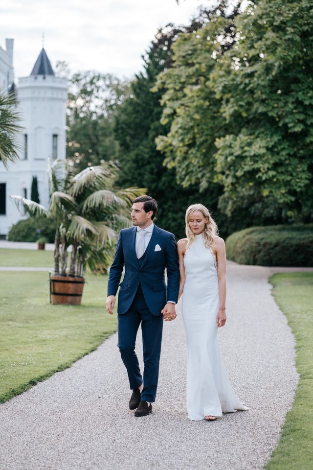 A couple walk hand in hand away from the camera along a gravel path at a wedding venue.