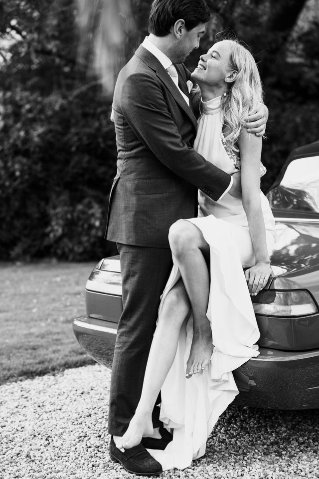 A couple ride laughing in a red classic Alfa Romeo convertible through a garden setting.