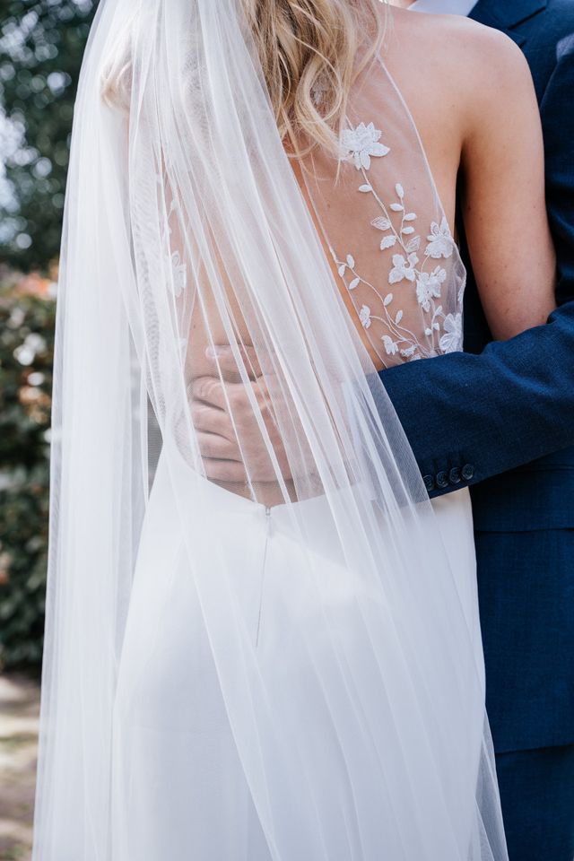A bride with long styled hair and a flowing veil embraces the groom, seen from behind.