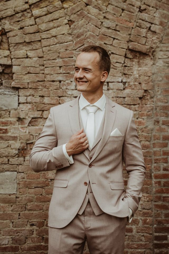 Bruidegom in crème pak staat zelfverzekerd tegen een verweerde stenen muur.