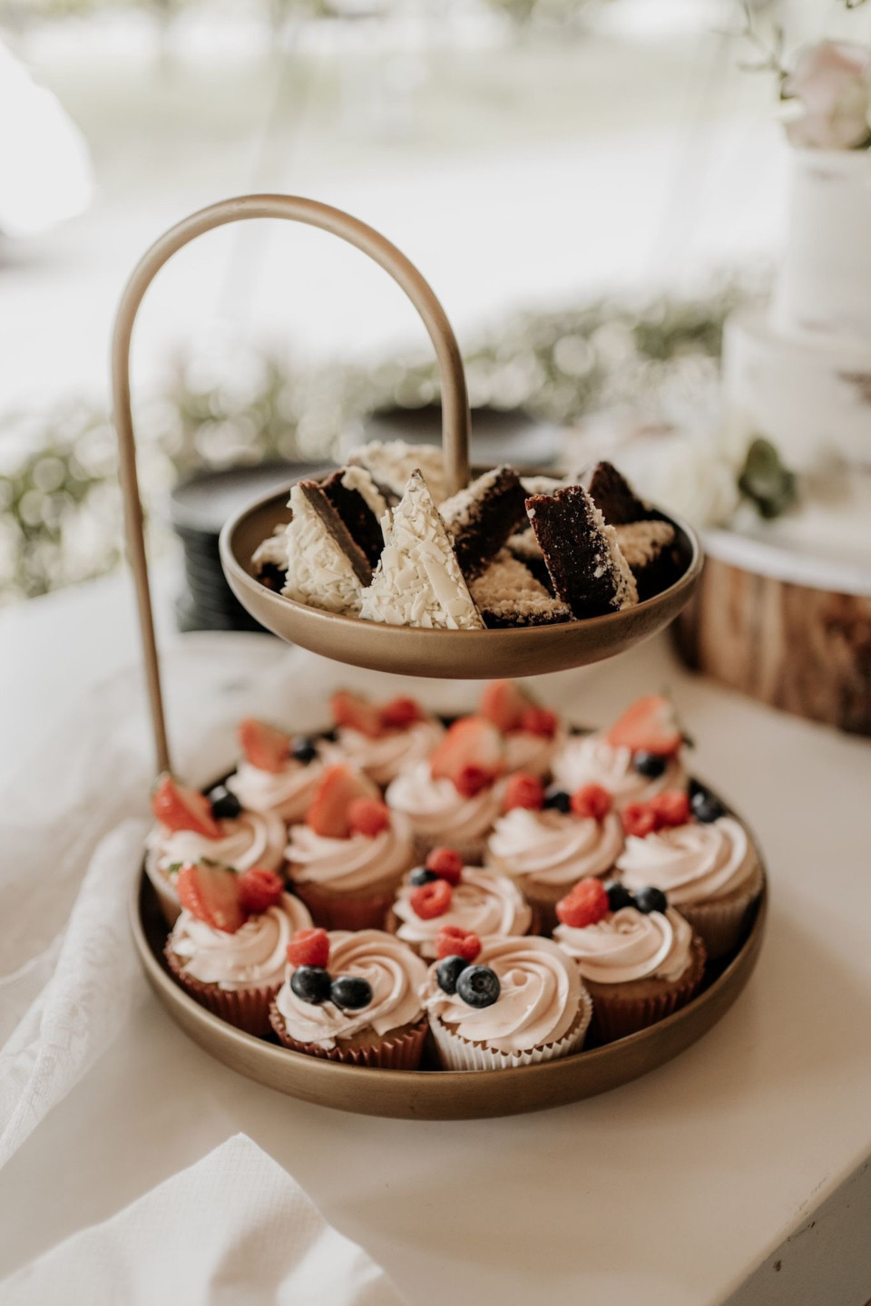 twee etagères met cupcakes met glazuur en fruit erop op een tafel voor een raam
