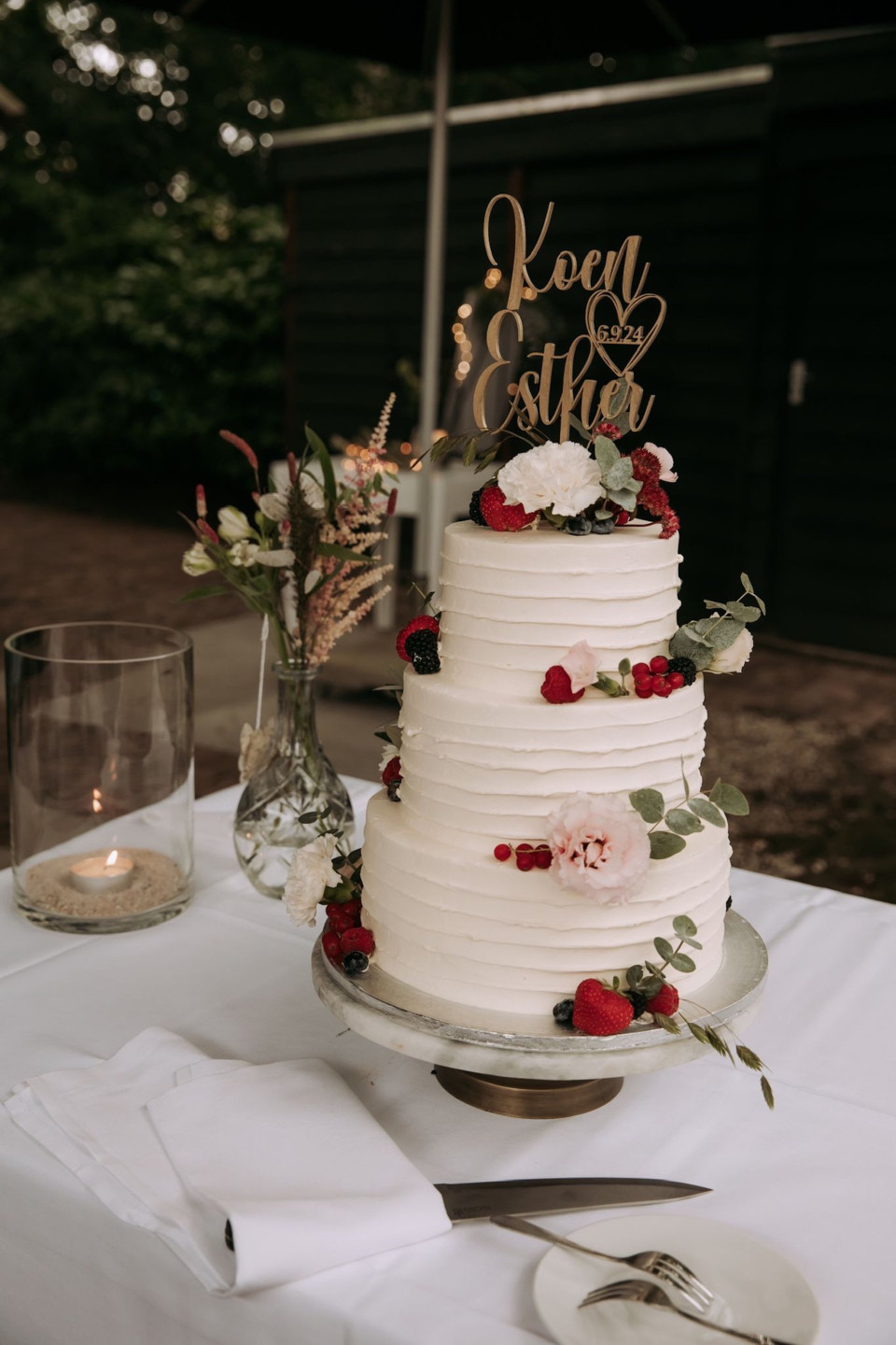 een bruidstaart op een tafel met een kaars en een glas wijn op de voorgrond en kaarslicht op de achtergrond
