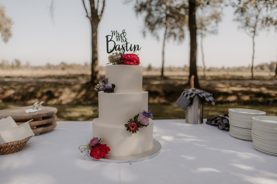 een bruidstaart op een tafel met borden en servetten op de voorgrond en bomen op de achtergrond