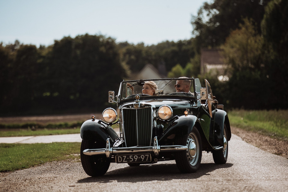 een paar mensen in een vintage auto die over een zandweg rijden met bomen op de achtergrond en gras op de voorgrond