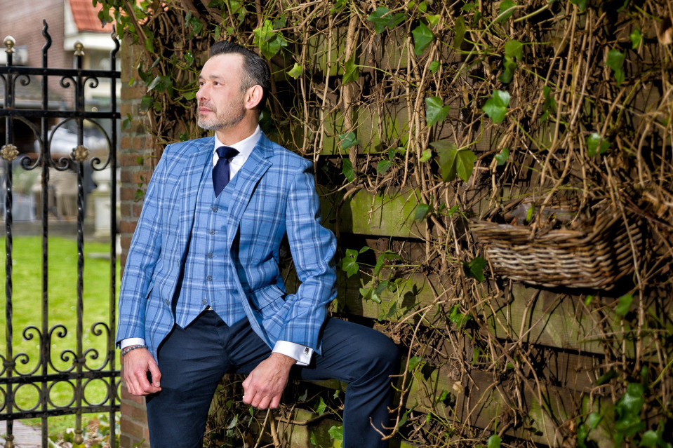 een man in een blauw pak en stropdas die tegen een hek leunt